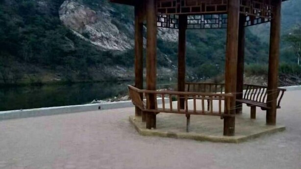 A wooden gazebo with a bench, surrounded by a paved area with a view of a river and mountains in the background.