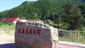 A large rock with Chinese characters, a building with a red roof, and a mountainous landscape in the background.
