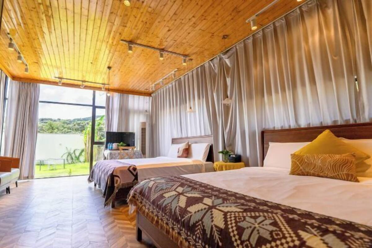 A hotel room with a wooden ceiling, two beds, a television, and a view of greenery outside.