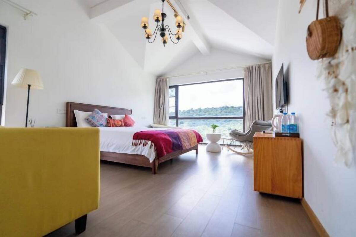 A bedroom with a bed, a desk, a chair, a lamp, a TV, and a view of the outdoors.