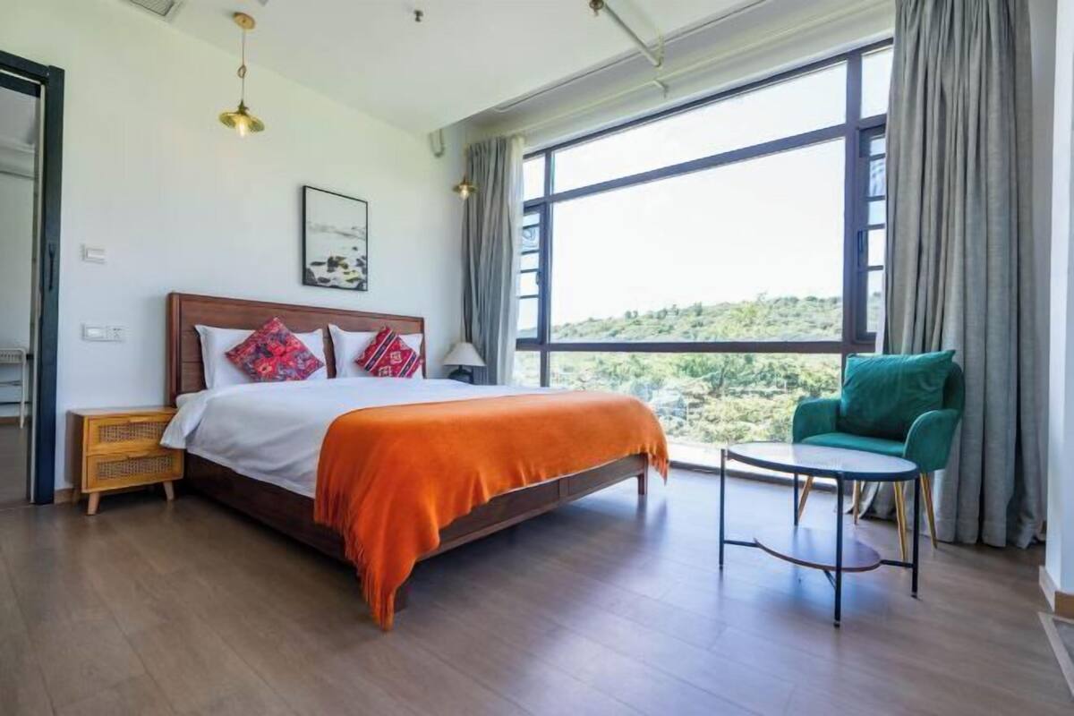 A bedroom with a large bed, a green chair, a small table, and a view of the outdoors through a large window.