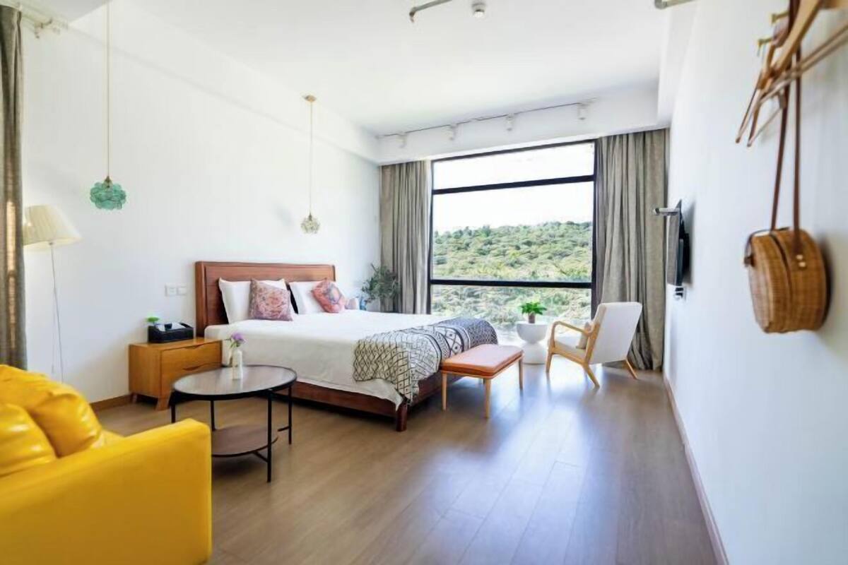 A modern hotel room with a large bed, a yellow armchair, a small round table, and a view of a green landscape through a large window.
