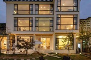 Un edificio de varios pisos con grandes ventanales, un acceso cubierto y mesas al aire libre.