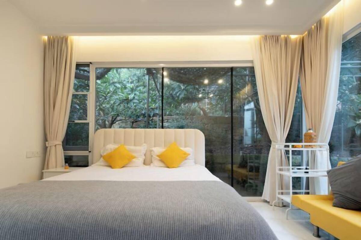 A bedroom with a large bed, beige curtains, and a view of greenery outside.
