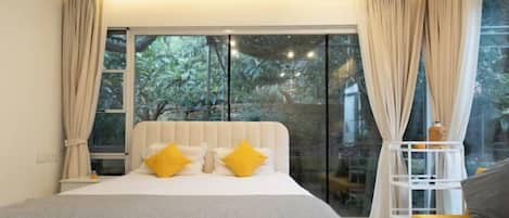 Un dormitorio con una cama grande, cortinas color beige y vistas al exterior con vegetación.