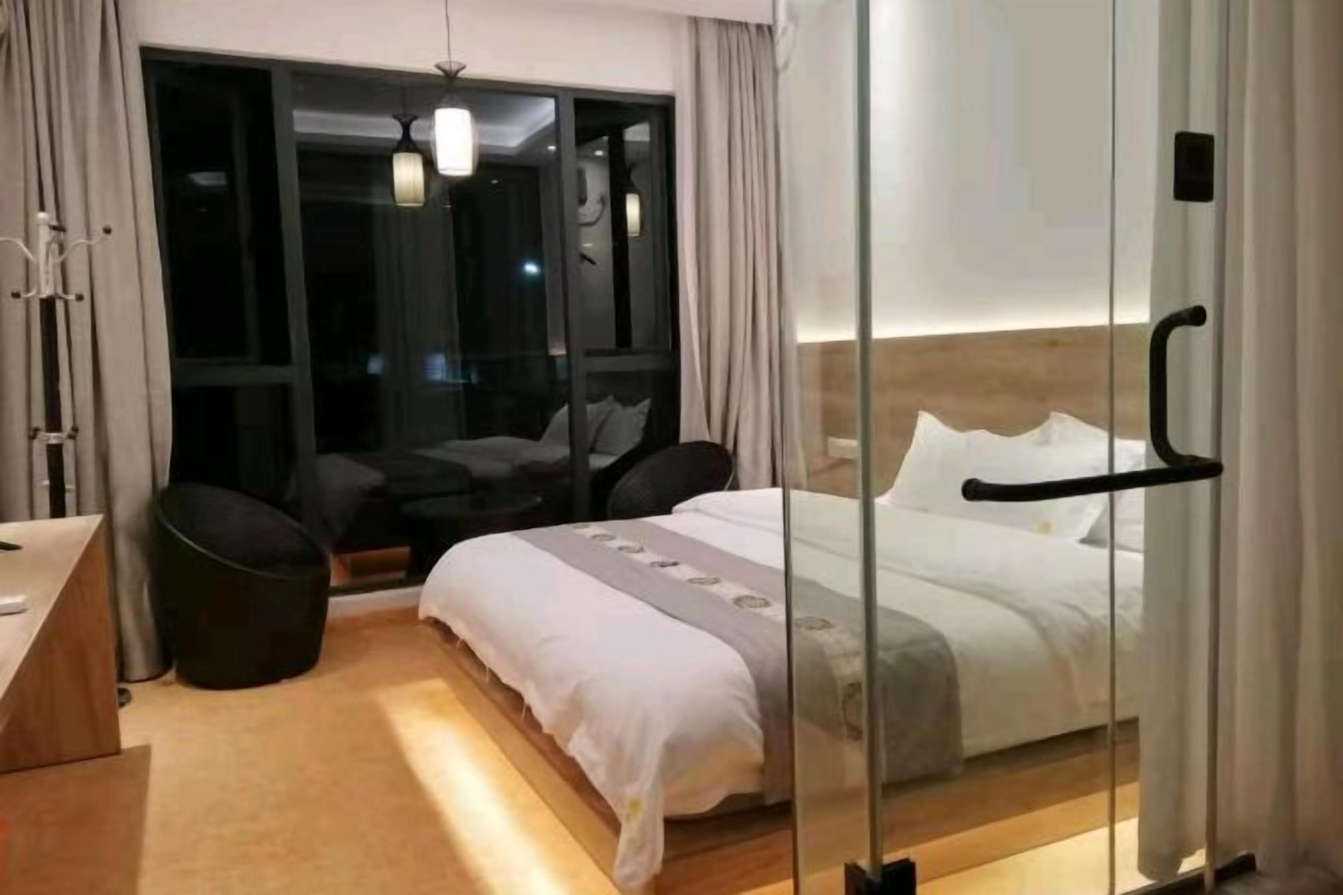 Una habitación de hotel moderna con un separador de vidrio, una cama con ropa blanca, un pequeño rincón de descanso con una silla y un escritorio.