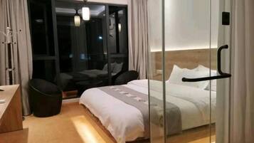 Una habitación de hotel moderna con un separador de vidrio, una cama con ropa blanca, un pequeño rincón de descanso con una silla y un escritorio.