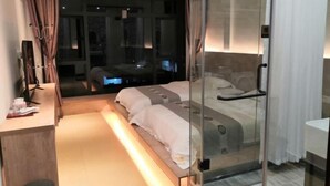 Une chambre moderne avec une cloison en verre, un lit avec une literie blanche, un bureau avec un ordinateur et une vue sur la ville la nuit.