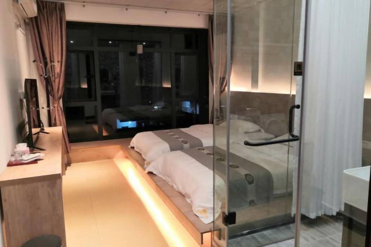 Une chambre moderne avec une cloison en verre, un lit avec une literie blanche, un bureau avec un ordinateur et une vue sur la ville la nuit.