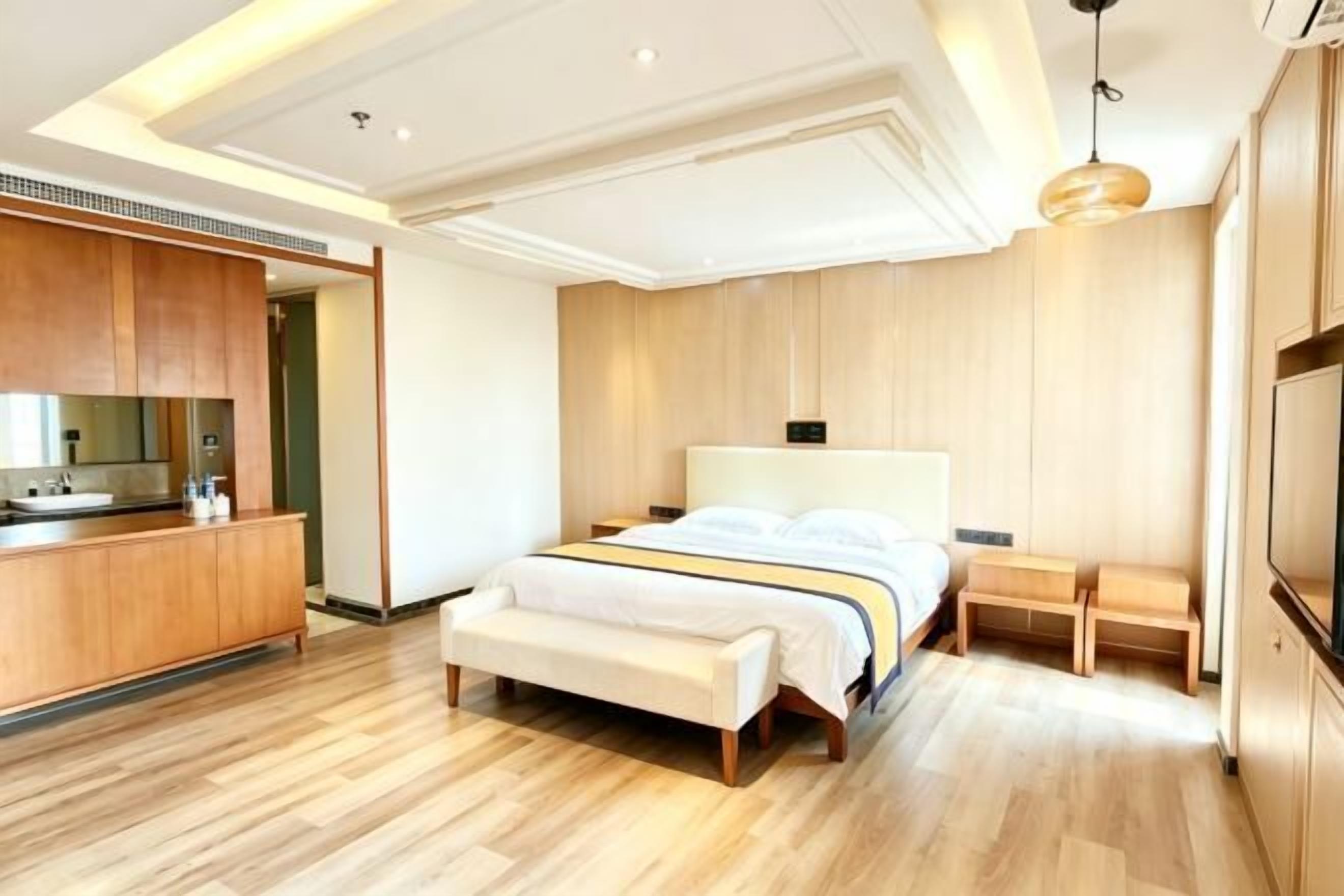 Ein modernes Hotelzimmer mit Holzboden, einem Bett mit weißem Kopfteil, einer Bank, einem kleinen Nachttisch und einem integrierten Fernseher.
