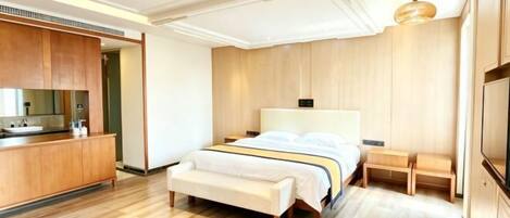 Ein modernes Hotelzimmer mit Holzboden, einem Bett mit weißem Kopfteil, einer Bank, einem kleinen Nachttisch und einem integrierten Fernseher.