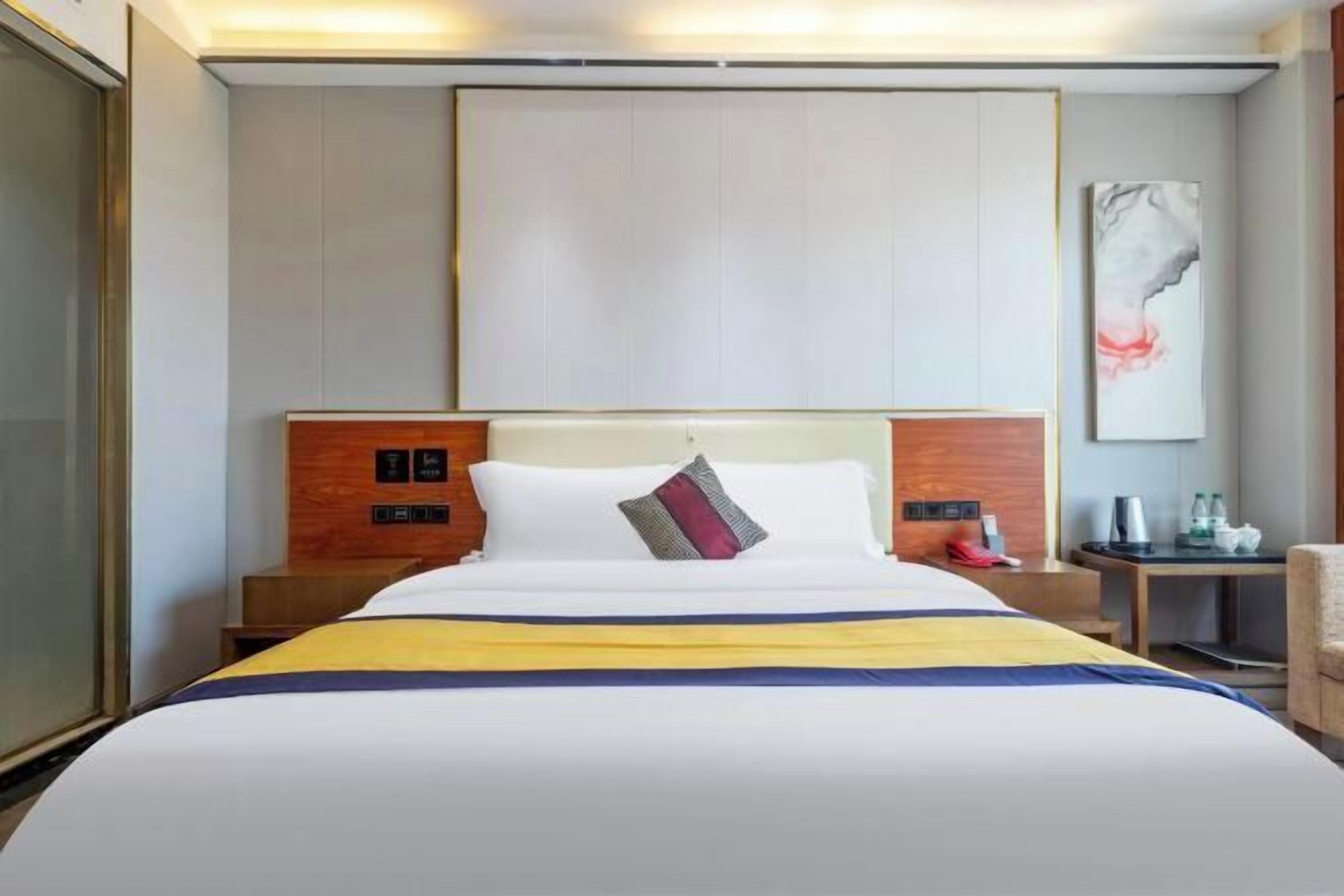A hotel room with a bed, bedside tables, a nightstand, a wall-mounted artwork, and a door.