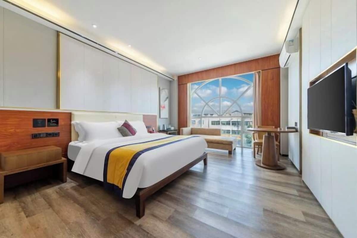 A modern hotel room with a large bed, a seating area, a TV, and a view of the cityscape.