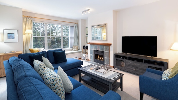 A living room with a blue sofa set, a fireplace, a flat-screen TV, and a large window with curtains. - Deer Run by Whistler Premier (Whistler)