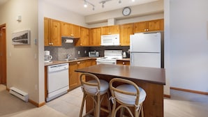 A modern kitchen with wooden cabinets, a central island with bar stools, and essential appliances like a microwave, oven, and refrigerator. - Deer Run by Whistler Premier (Whistler)