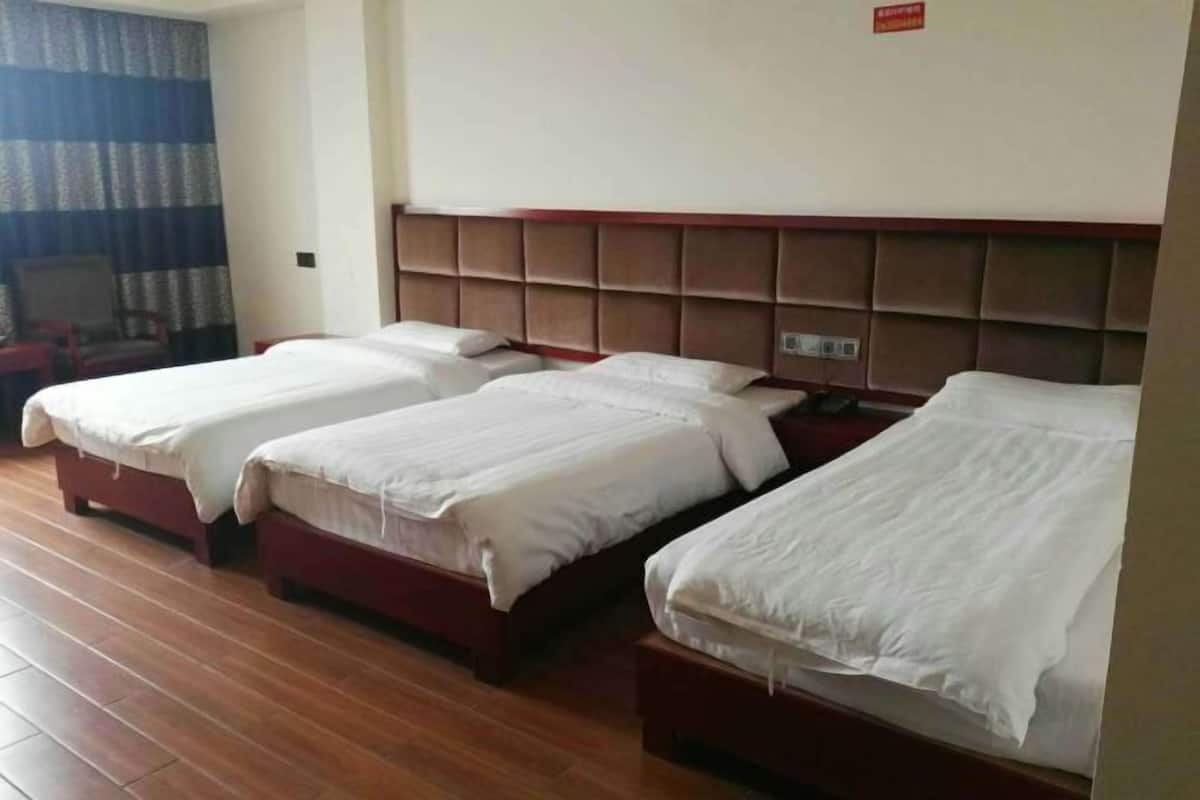 A hotel room with three single beds aligned against a wall, wooden flooring, and a window with curtains.