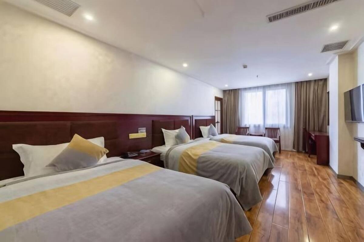 Habitación de hotel con tres camas, suelo de madera, televisor y una ventana con cortinas.