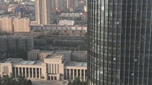 Un edificio alto con fachada de vidrio moderno, rodeado de edificios de altura media y una plaza central con algunos árboles.