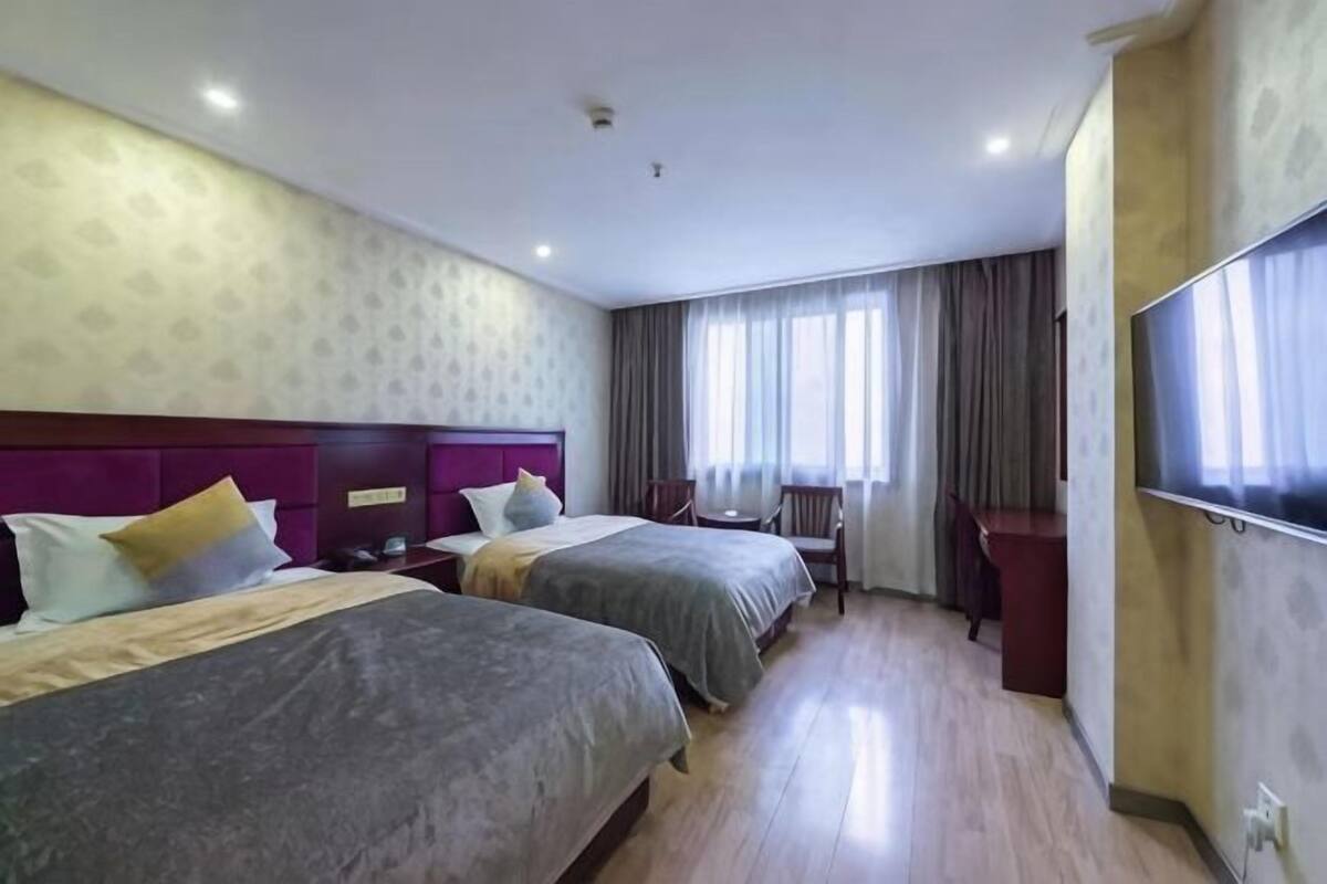Habitación de hotel con dos camas, un escritorio, una silla y un gran ventanal con cortinas.