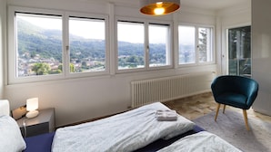 A bedroom with a bed, a chair, a nightstand, a lamp, and a view of a town through large windows.