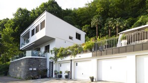 A modern two-story house with a balcony, garage doors, and a stone wall.