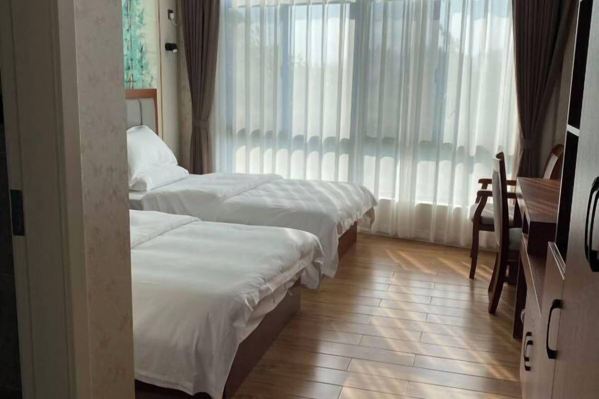 A hotel room with two beds, a wooden floor, a desk with a chair, and a large window with curtains.