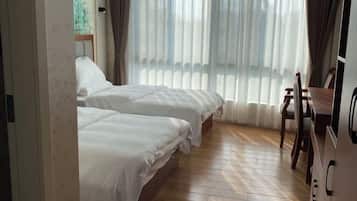 A hotel room with two beds, a wooden floor, a desk with a chair, and a large window with curtains.