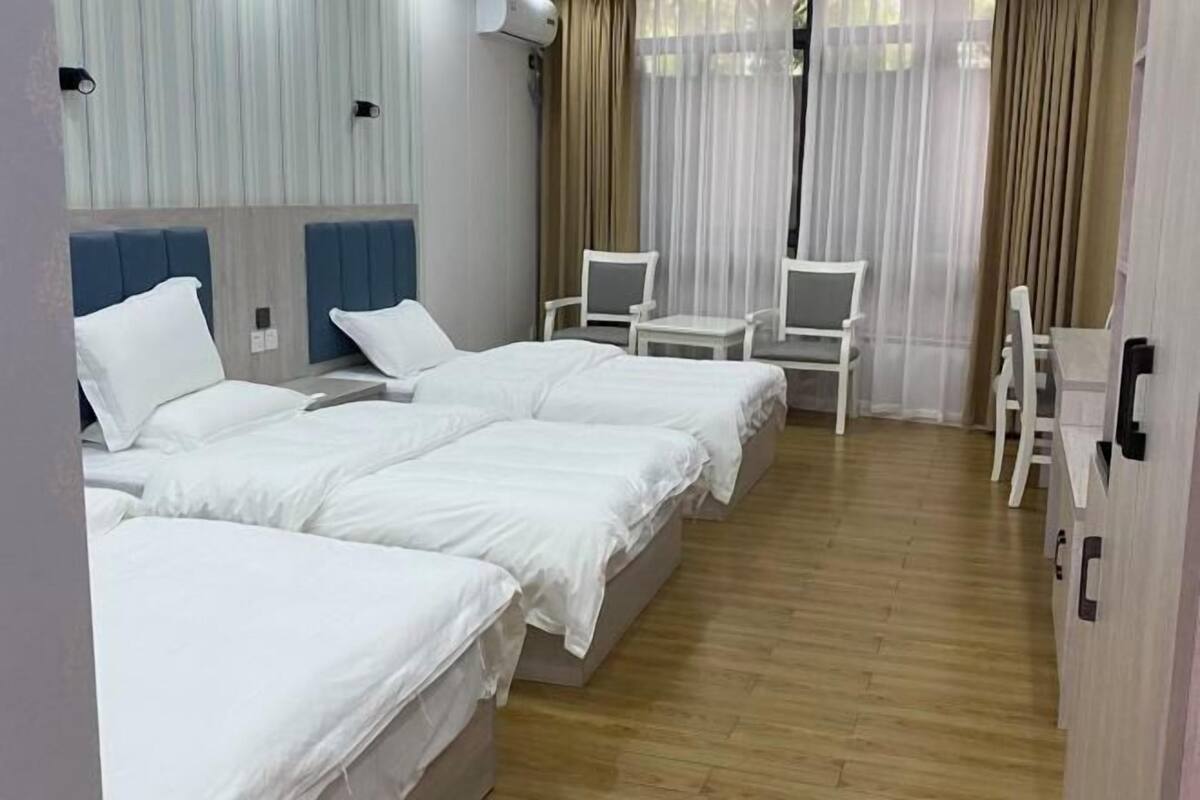 A hotel room with three beds, a small table, two chairs, and a window with sheer curtains.