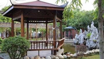 Um gazebo de madeira com telhado, rodeado por um lago com estátuas brancas, e uma pequena ponte que leva a um edifício.