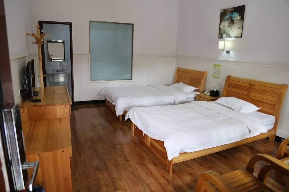 A hotel room with two wooden beds, a wooden desk, a mirror, a window with blinds, and a wall-mounted light fixture.