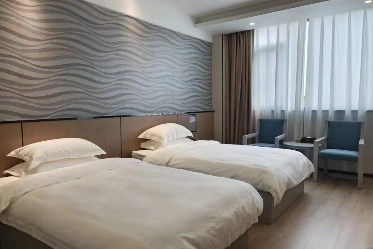Quarto de hotel com duas camas, cabeceira com design ondulado, uma mesinha, duas cadeiras e janela com cortinas.