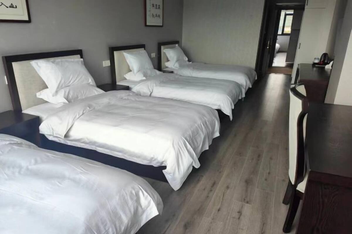 Quarto de hotel com três camas, piso de madeira e uma escrivaninha com cadeira.