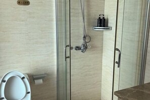 A bathroom with a toilet, a shower area with a glass door, and a shelf with toiletries. - Baolong Furui Hotel (Changzhou)