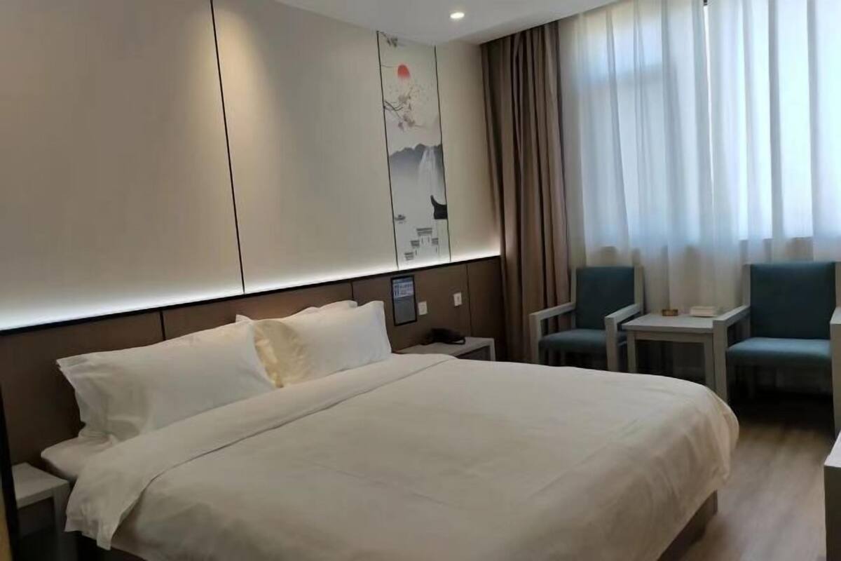 Quarto de hotel com uma cama grande, uma mesinha de cabeceira, uma cadeira, uma pequena mesa e um painel decorativo na parede.