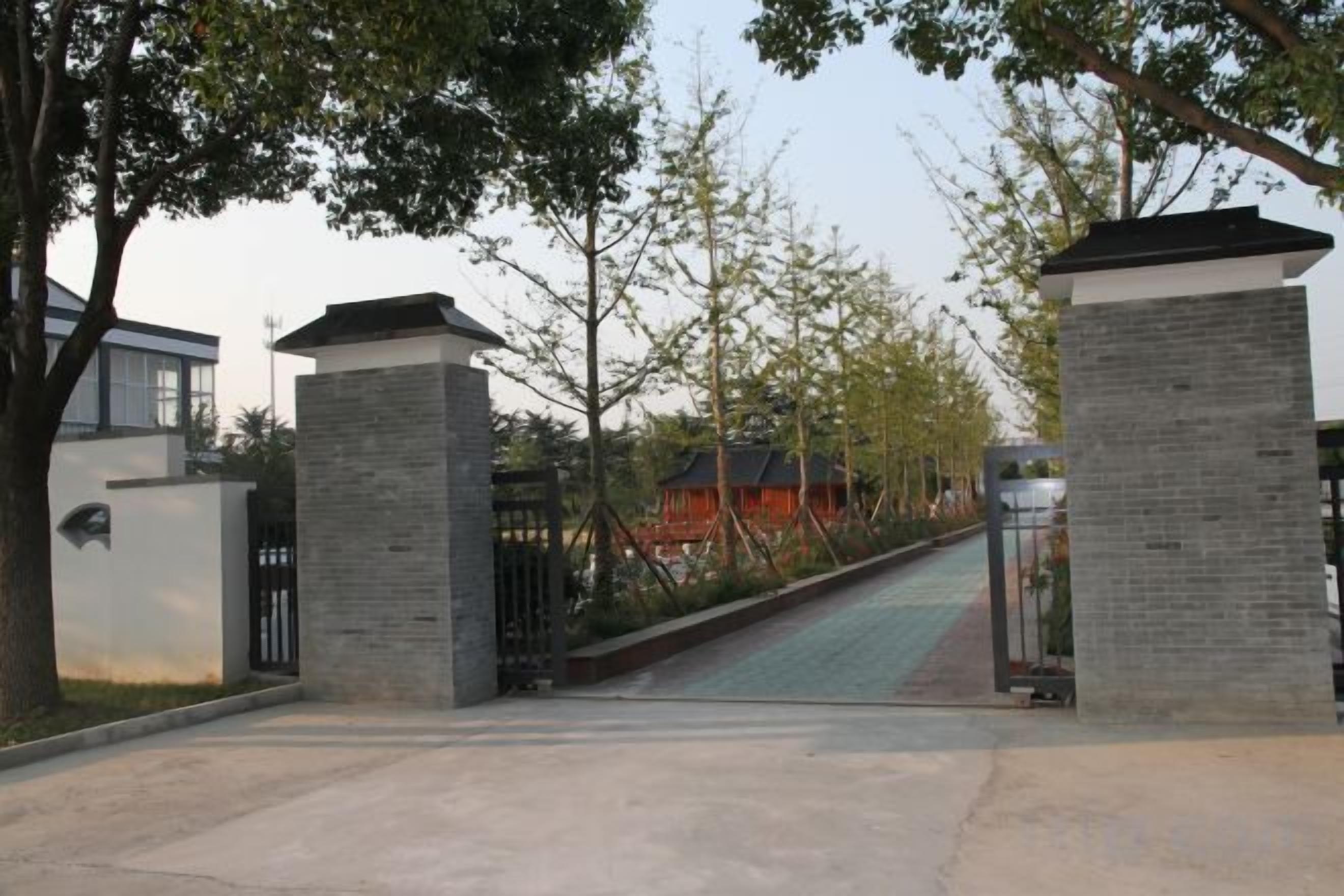 A gated entrance with a pathway leading to a building, surrounded by trees and a clear sky.