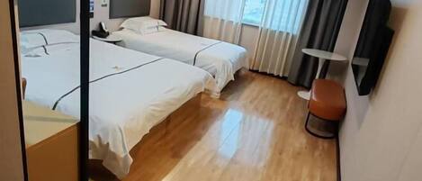 A hotel room with two beds, a bedside table, a chair, a small table, and a window with curtains.