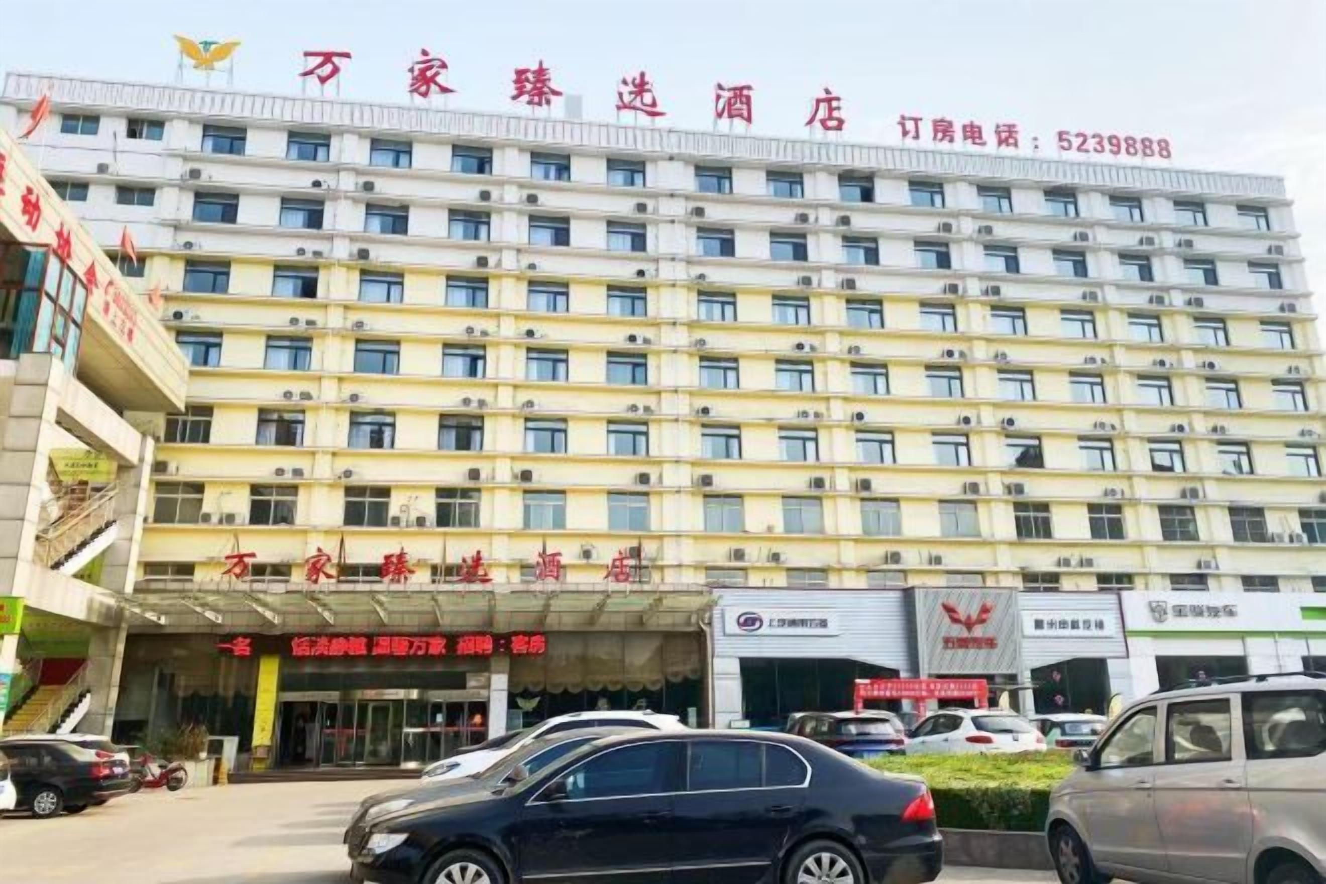 Un hotel de varios pisos con una fila de ventanas en la fachada, un cartel con el nombre del hotel y un estacionamiento con coches.