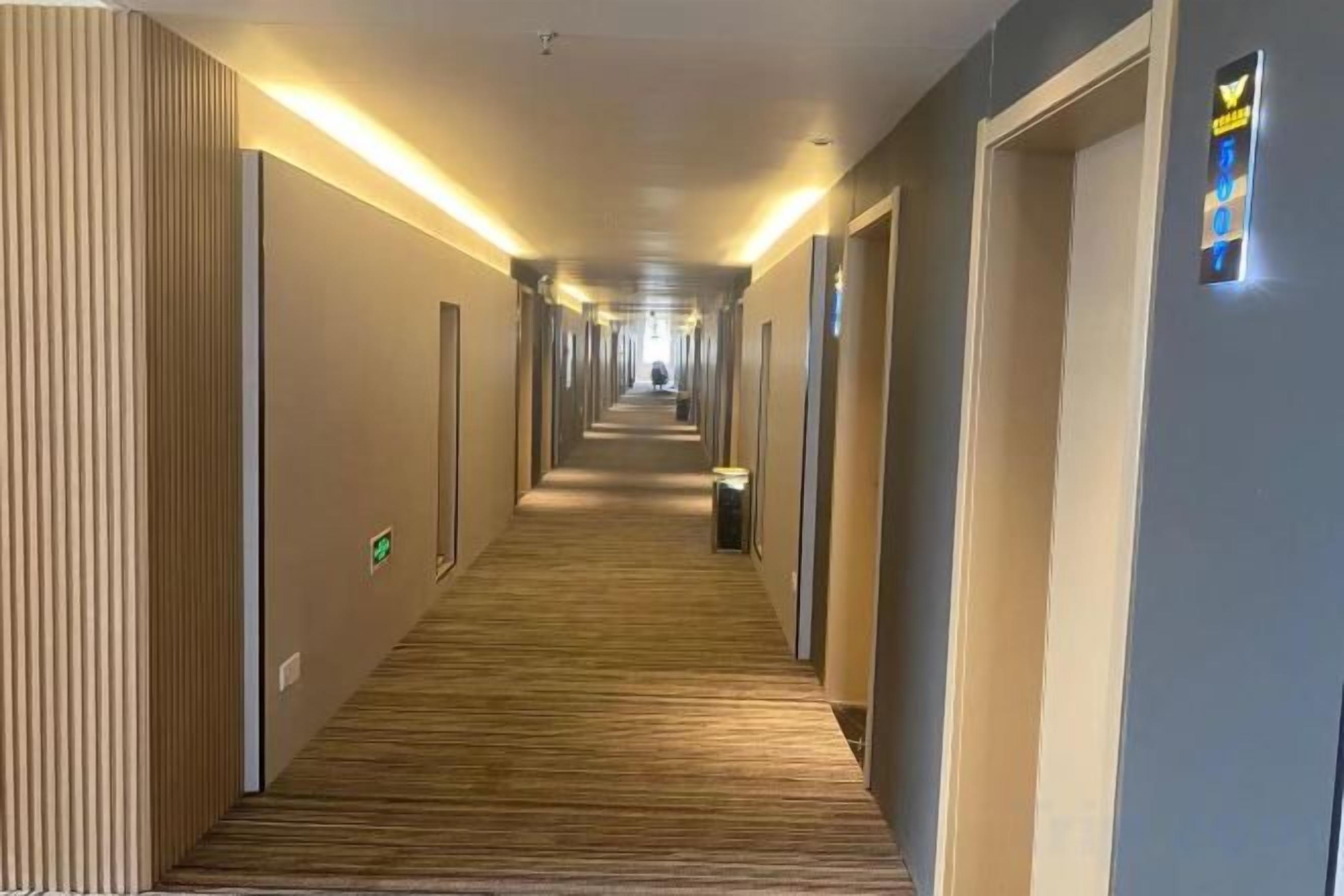 A hotel corridor with a wooden floor, doors on both sides, and a lighted sign on the right wall.