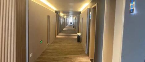 A hotel corridor with a wooden floor, doors on both sides, and a lighted sign on the right wall.