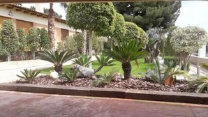 Ein angelegter Garten mit verschiedenen Pflanzen und Bäumen, einem gepflasterten Weg und einem Gebäude im Hintergrund.
