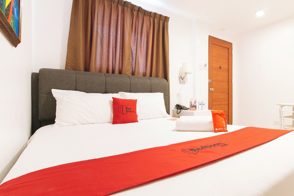 A hotel room with a large bed, a grey headboard, white bedding, a red pillow, a red blanket, a wooden door, a white chair, and a painting on the wall.