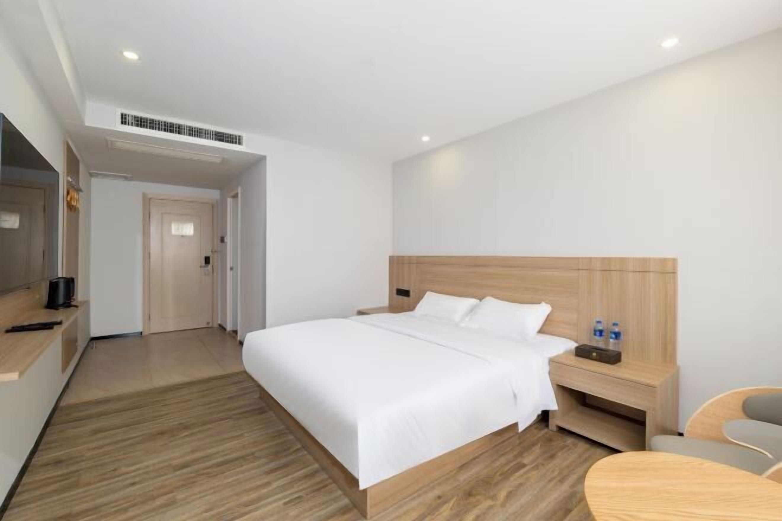 A hotel room with a wooden bed, a bedside table with water bottles, a desk with a computer, and a chair.
