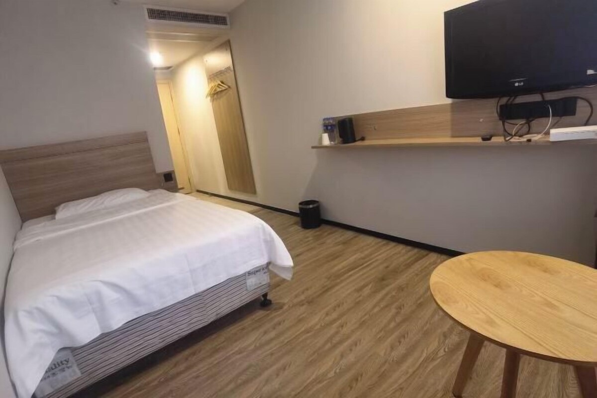 A hotel room with a bed, a small wooden table, a TV mounted on the wall, and a mirror above the bed.