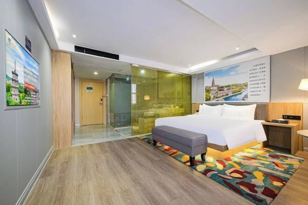 Une chambre d’hôtel moderne avec un grand lit, un petit bureau, une cloison en verre et un tapis aux couleurs vives.