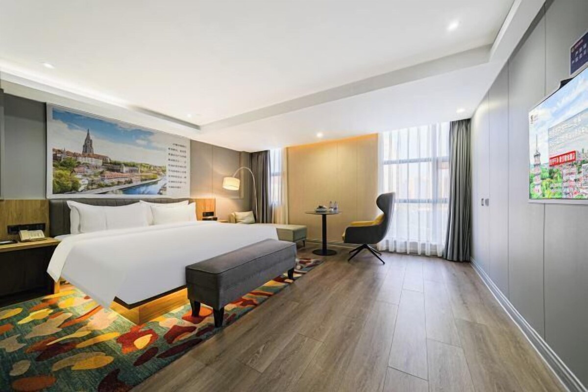A hotel room with a large bed, a desk, a chair, and a view of a cityscape through the window.