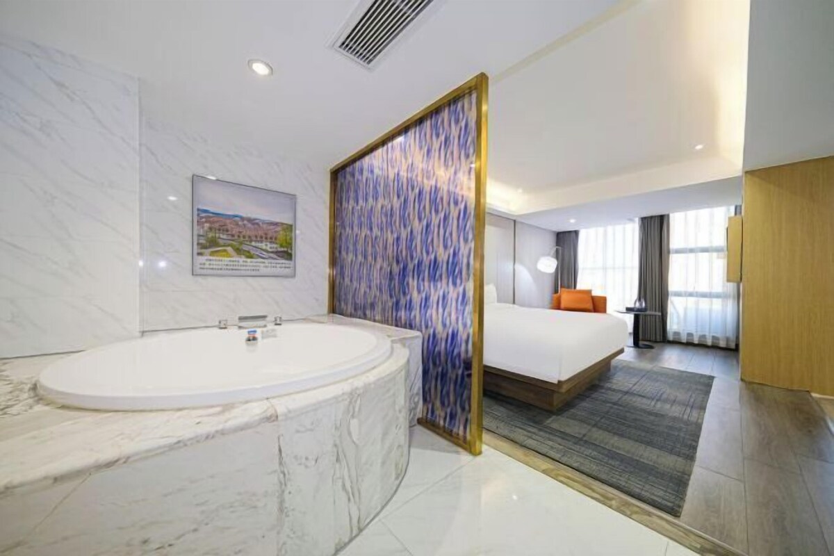 A modern hotel room with a marble bathtub, a large bed, and a view of the city.