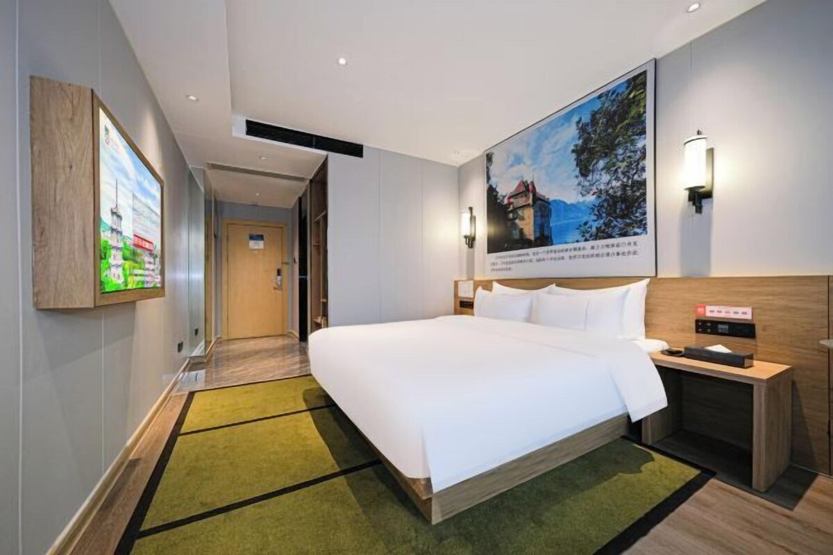 A hotel room with a large bed, a nightstand, a wall-mounted lamp, and a painting.