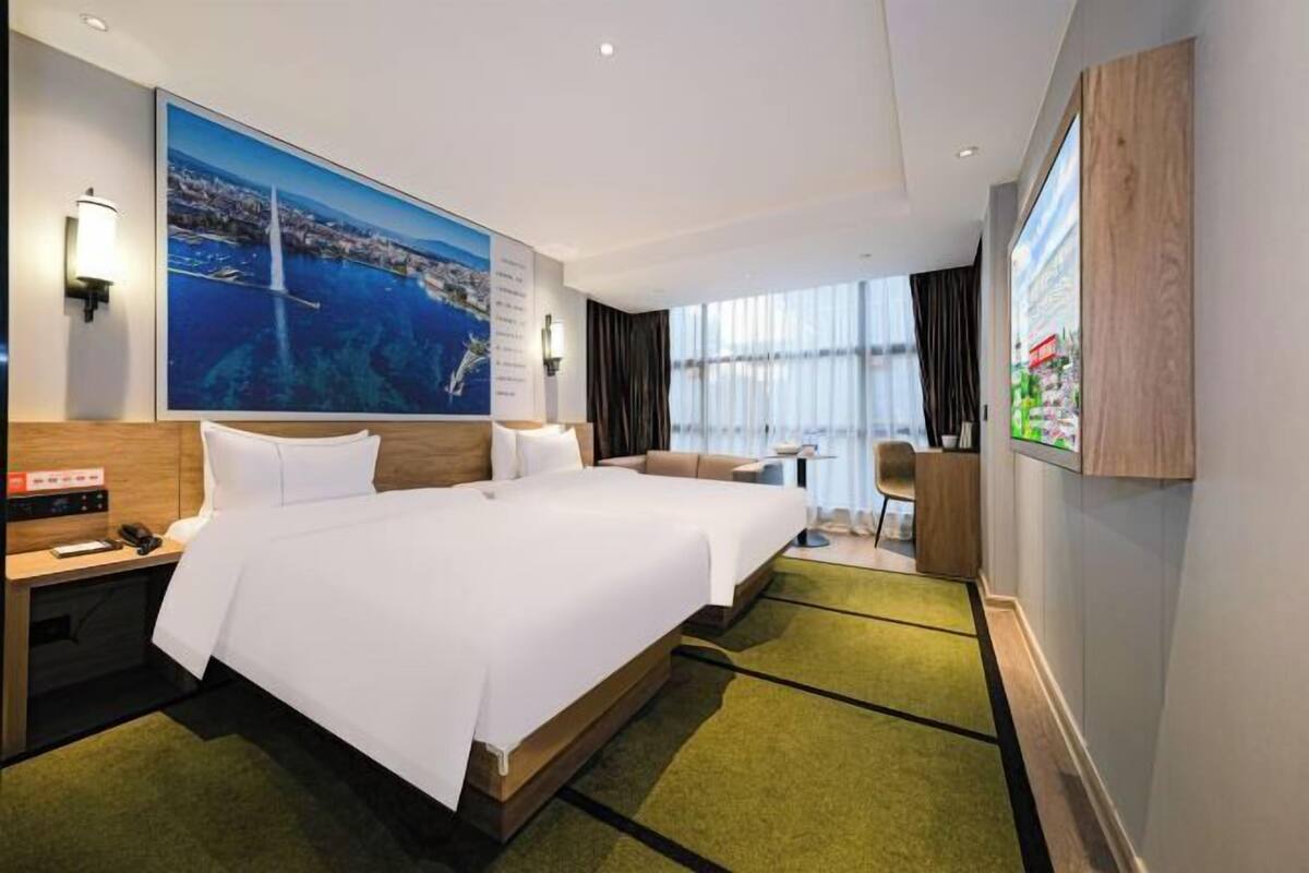Une chambre d’hôtel avec deux lits, une grande fenêtre, un tableau représentant un paysage côtier et un minibar.