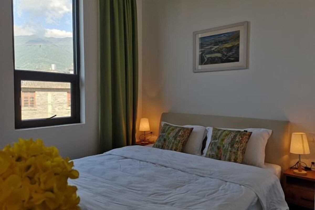 Habitación de hotel con cama, dos lámparas de noche, ventana con vistas y un cuadro en la pared.