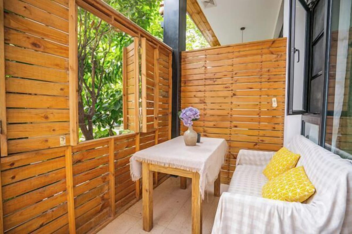 Una habitación acogedora con un muro de listones de madera, un sofá blanco con cojines amarillos y una pequeña mesa con un jarrón de flores.